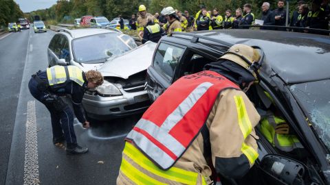 A member of the fire department examines a car involved in an accident. A policewoman marks the final position of another car involved in the accident with chalk. Other members of the police and fire department can be seen in the background, as well as vehicles.