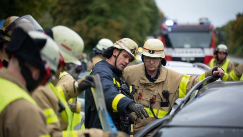 Several firefighters stand next to a car involved in an accident and confer. A fire engine can be seen in the background.
