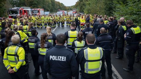A group of police and firefighters stand together in a circle on a closed highway after a traffic accident drill. One person speaks to the crowd. Several police and fire engines can be seen in the background.